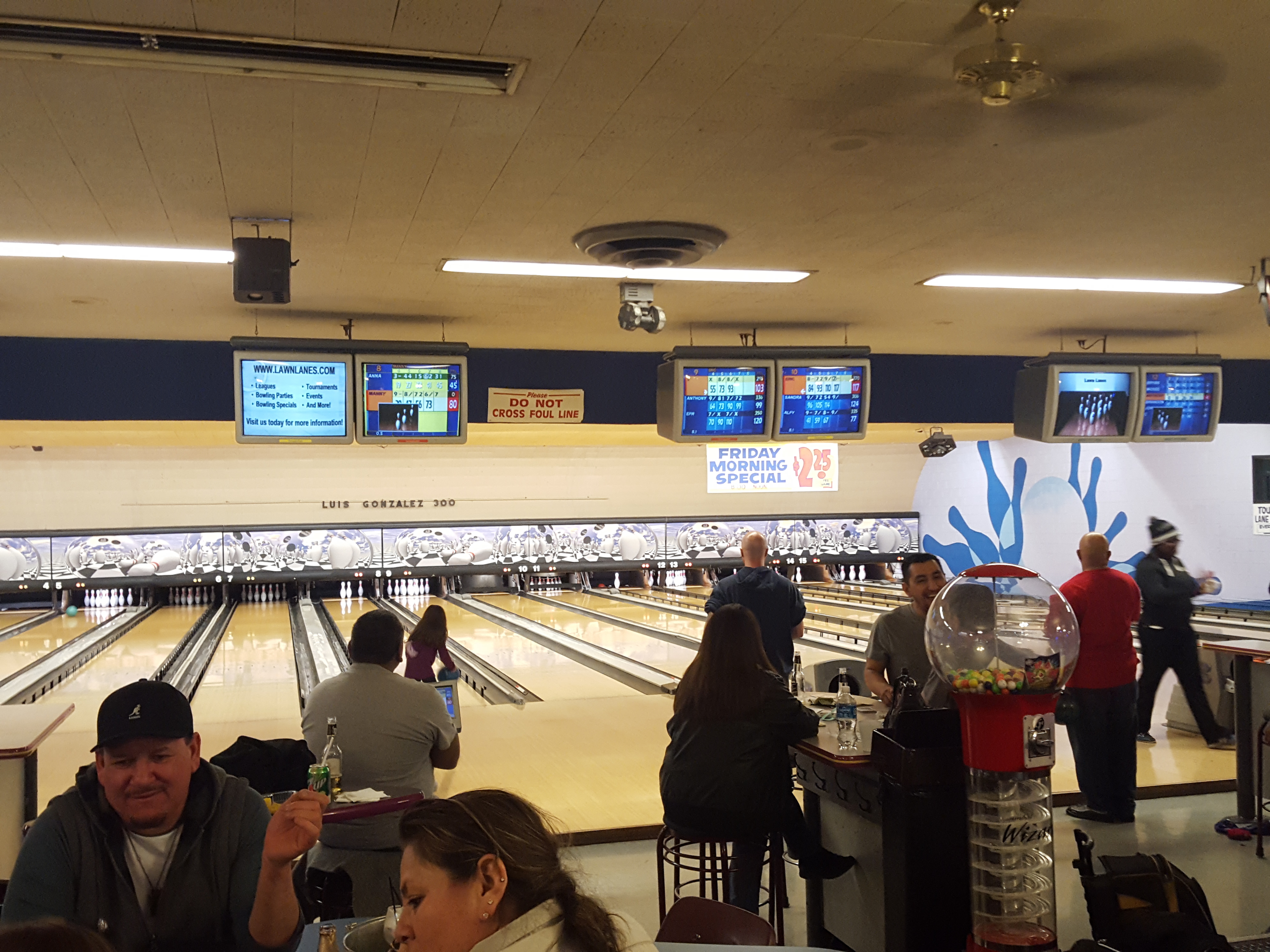 People bowling and socializing at a bowling center with scoring monitors and a Friday Morning Special sign visible.