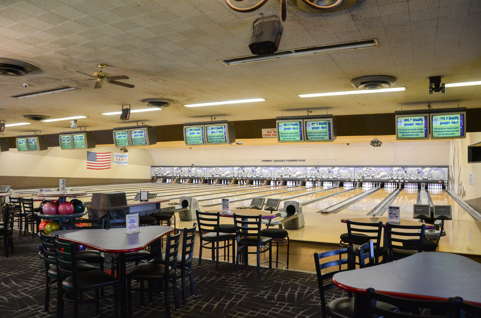 A wide view of several bowling lanes with dining tables in the foreground and a league sign on the back wall.