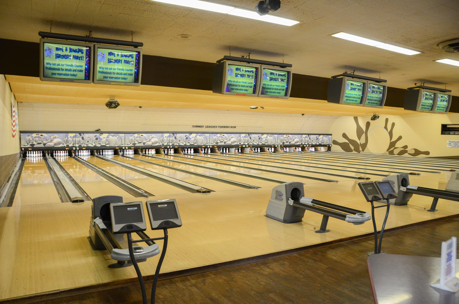A row of several well-lit bowling lanes with overhead monitors advertising birthday parties.