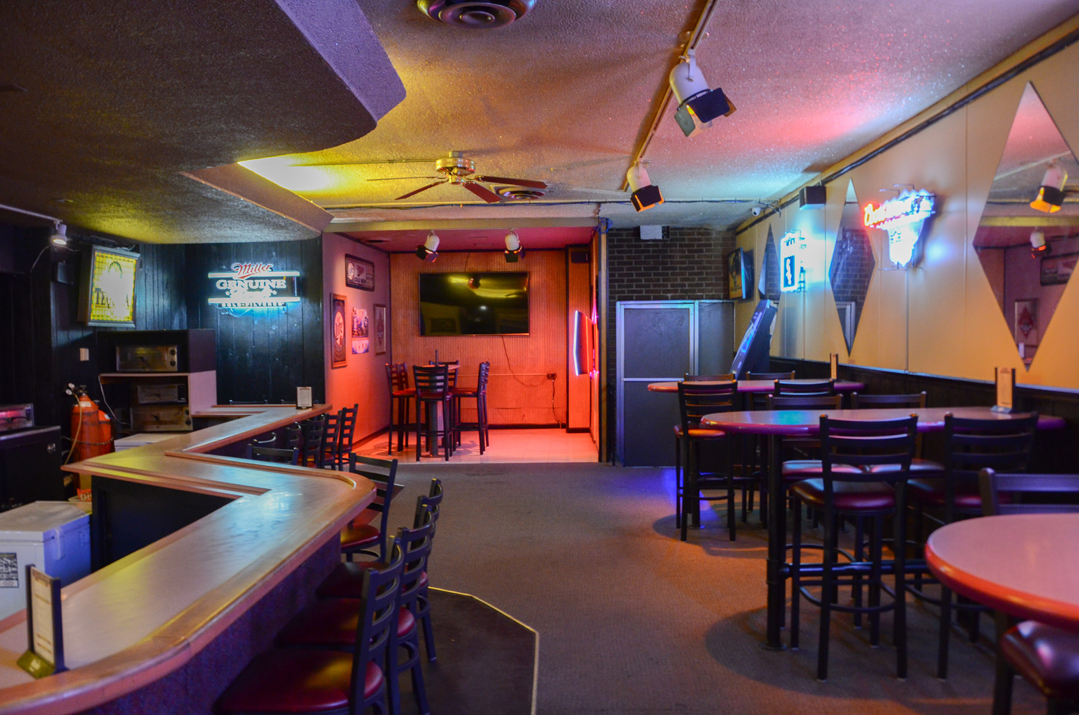 A bowling center lounge with a wooden bar, high-top tables, and neon beer signs under colorful ambient lighting.