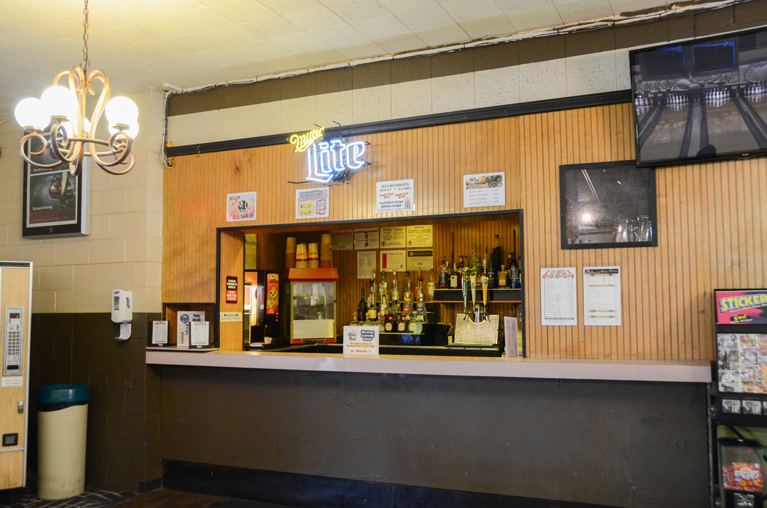 A wood-paneled bar counter with a neon beer sign, drink menus, and a TV showing bowling lanes.