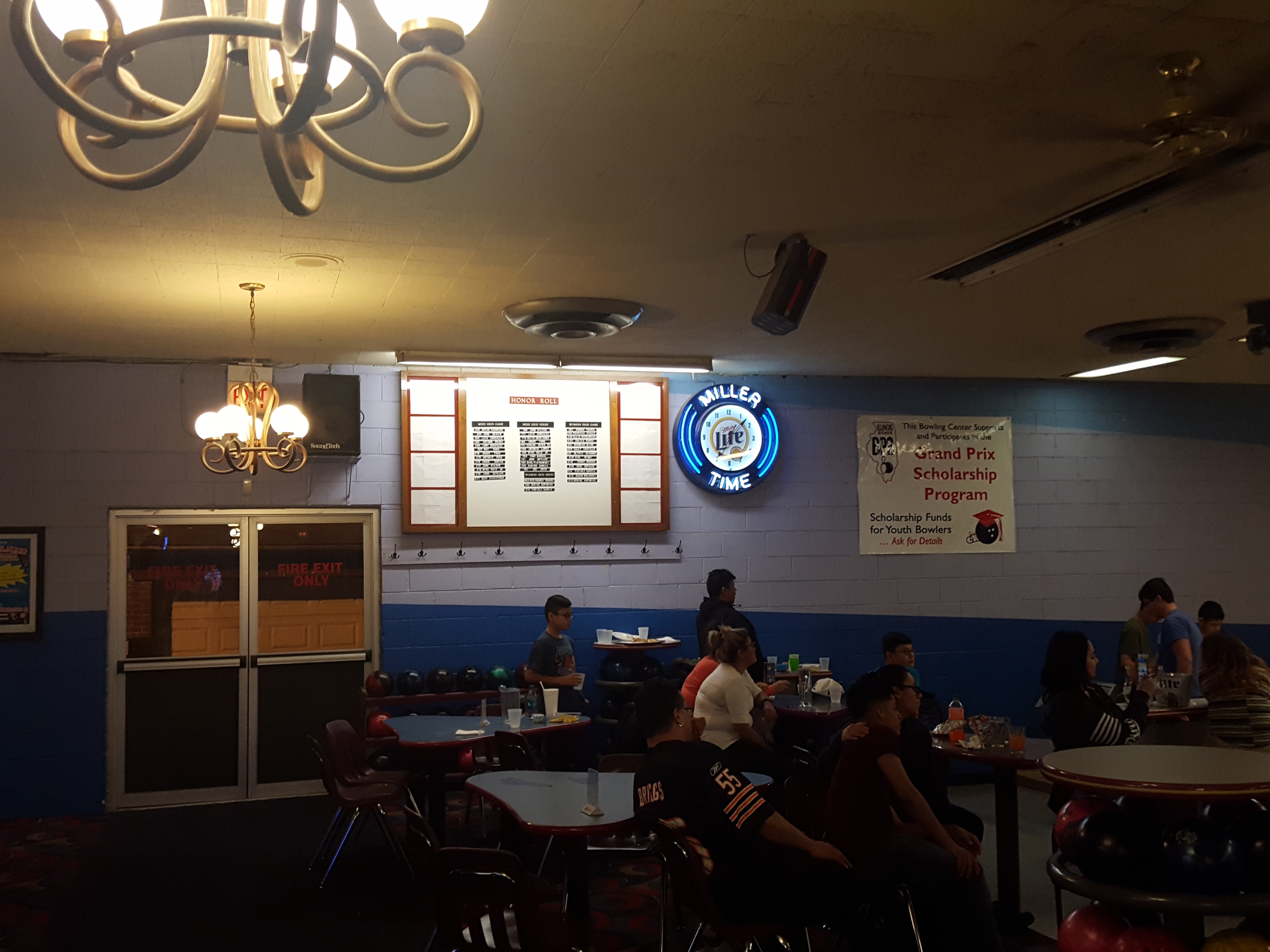 A social seating area in a bowling alley with an honor roll board, neon clock, and people at tables.