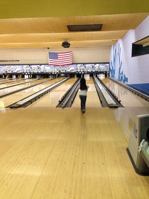A person standing on a bowling lane approach in front of an American flag.