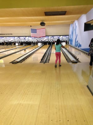 A young person standing on a bowling lane with bumpers up, facing pins beneath an American flag.