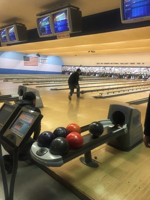 A man in a black jacket bowls on a wooden lane at a bowling center with a ball return in the foreground.