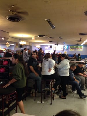 A group of people sitting on stools and chairs in a bowling alley concourse next to a ball rack.