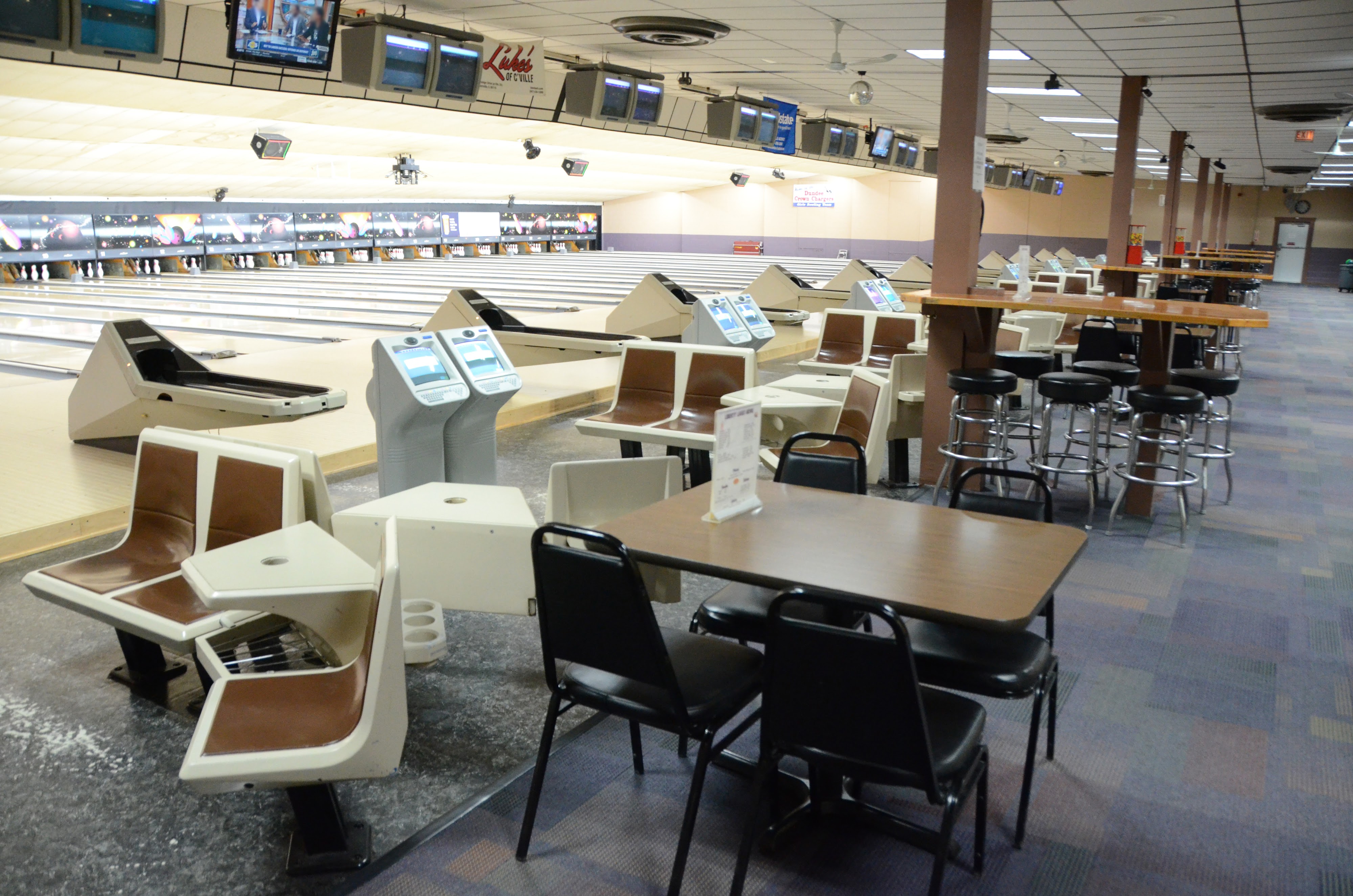 A clean bowling center interior showing multiple lanes, seating areas, scoring consoles, and overhead monitors.