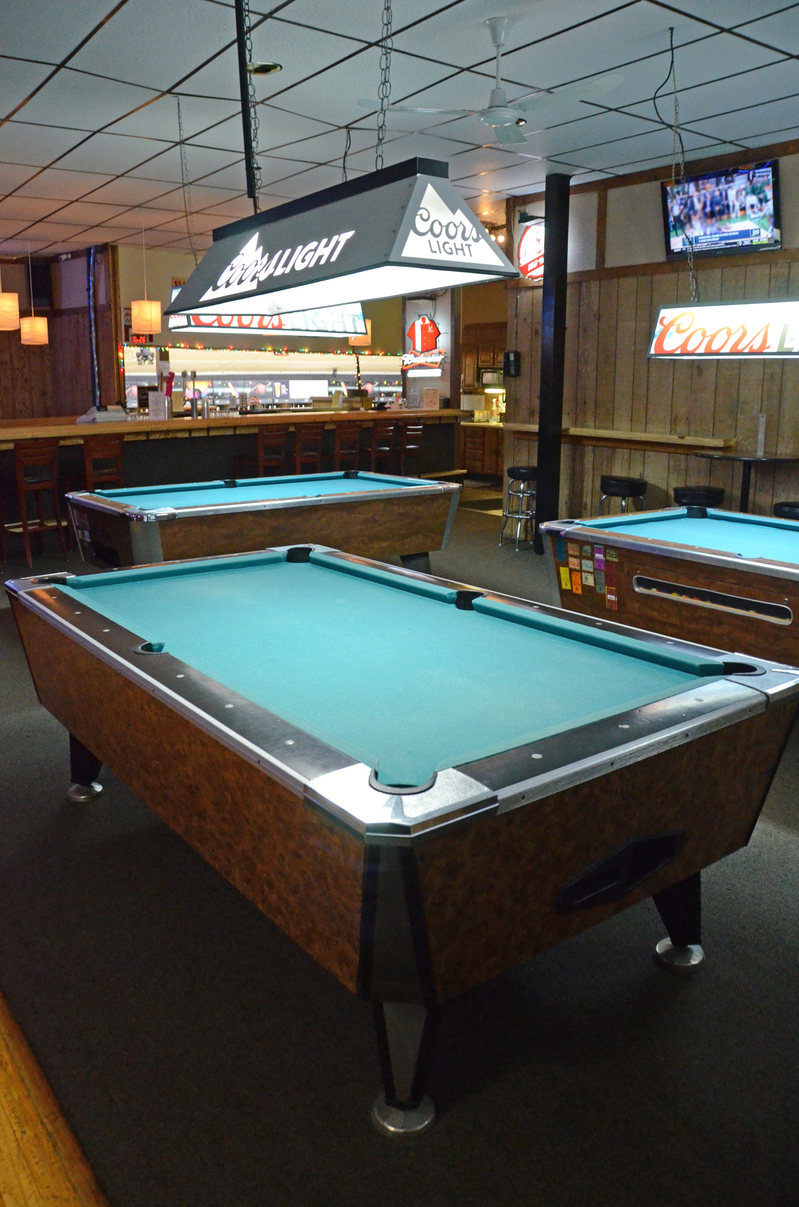 Three billiard tables with teal felt inside a wood-paneled room at a bowling center.