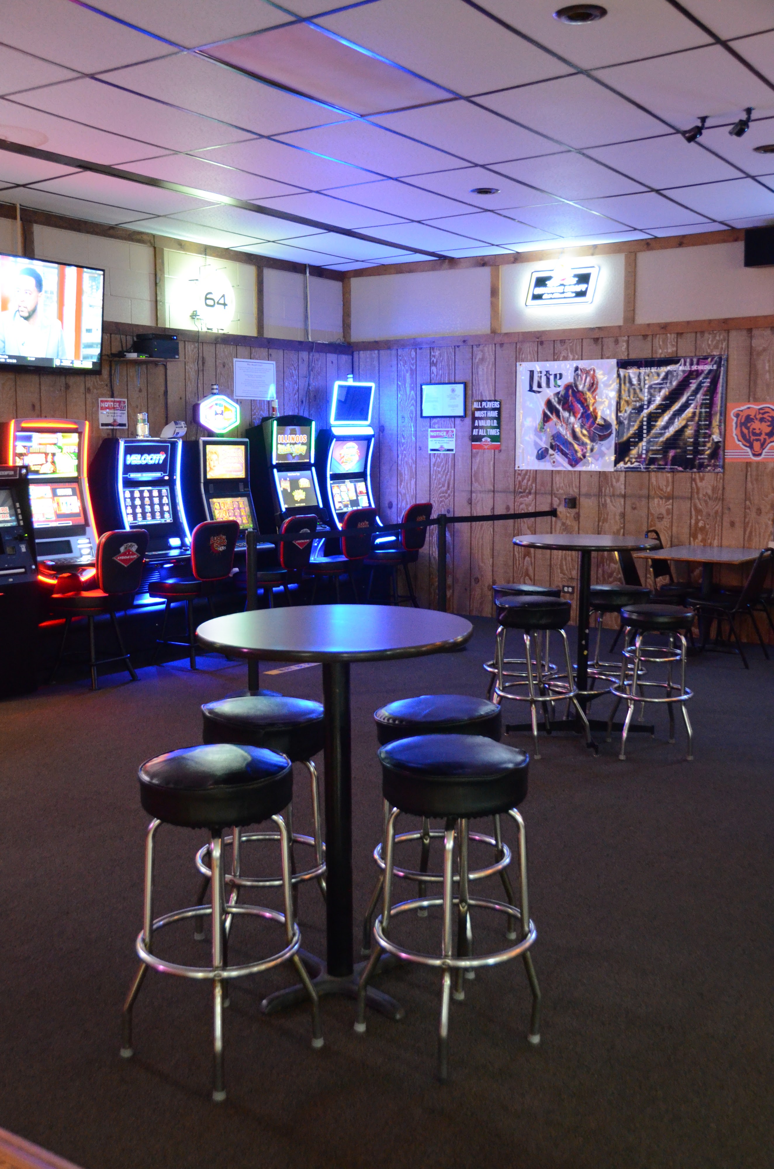 A lounge area with high-top tables, stools, and a row of lit-up video gaming machines against a wood wall.