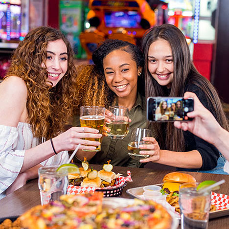Three women smile and hold up drinks for a photo while sitting at a table filled with pizza and burgers.