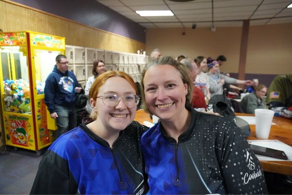 Two women in matching blue and black bowling jerseys smile for a photo inside a bowling center.