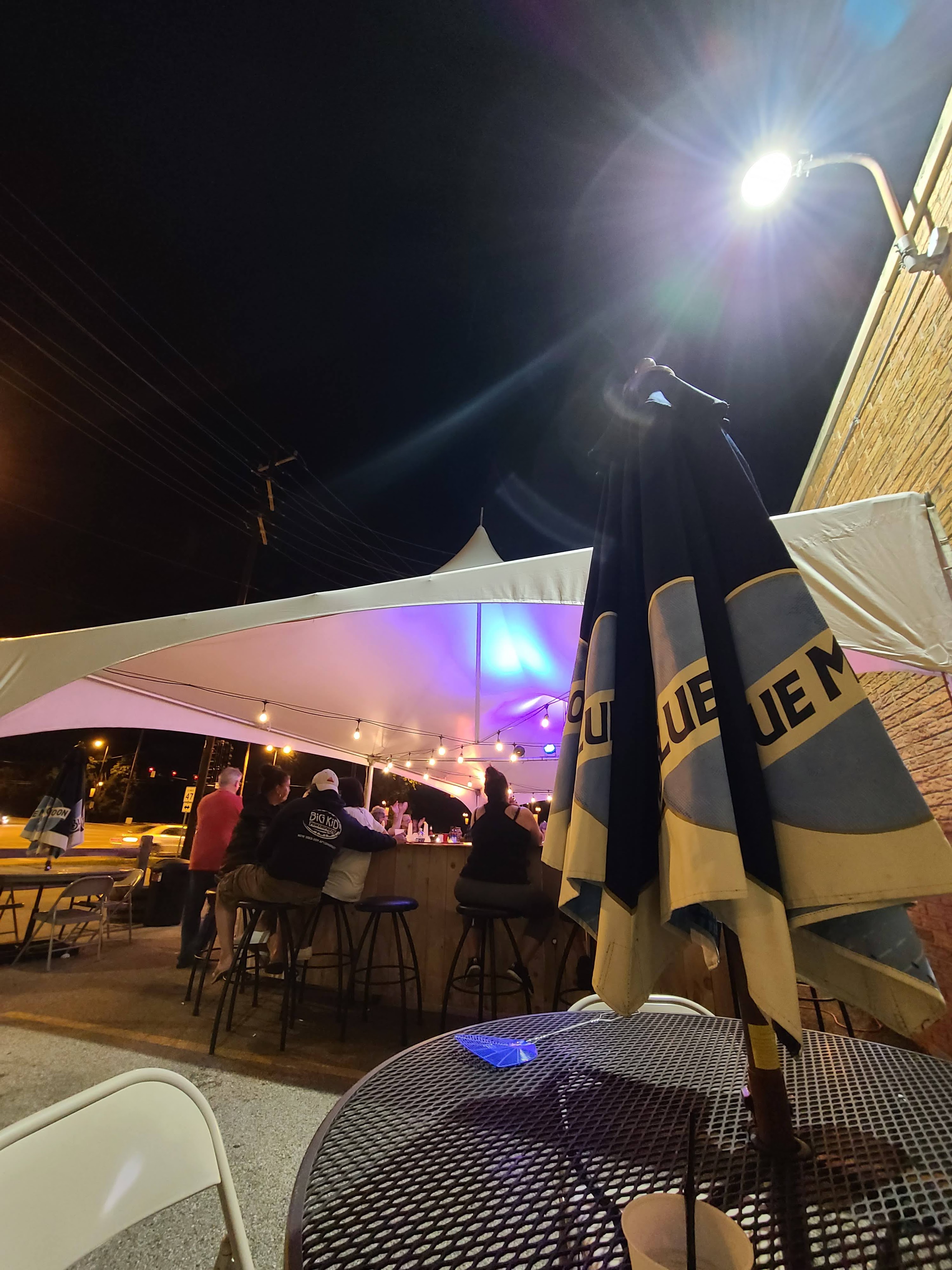 A nighttime outdoor patio bar with people sitting under a large white tent lit with purple and string lights.