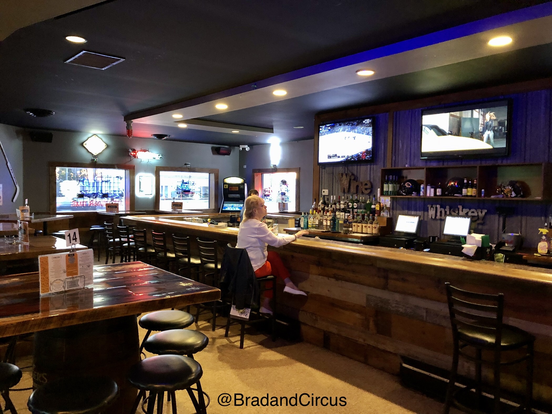 A woman sits at a long wooden bar with liquor bottles and televisions in a dimly lit lounge.