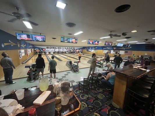A wide-angle interior view of Bowl-Hi Lanes with people bowling and patrons sitting at high-top bar tables.