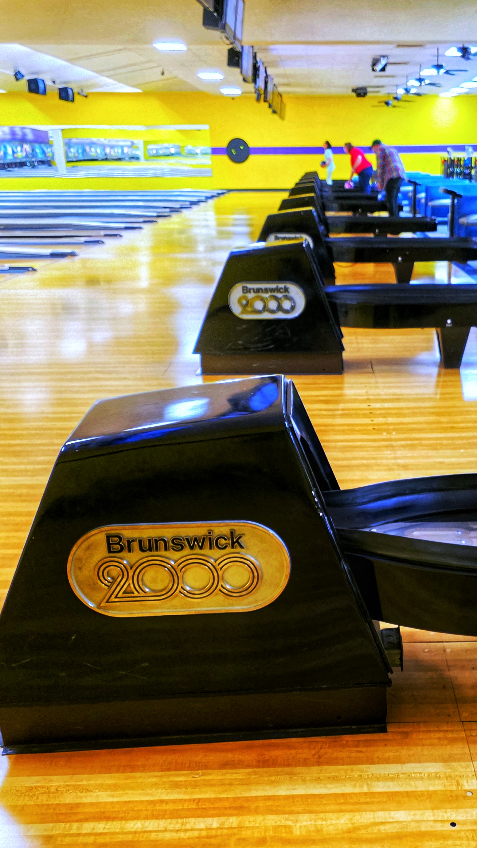 A row of black Brunswick 2000 ball returns on wooden bowling lanes with bowlers in the background.