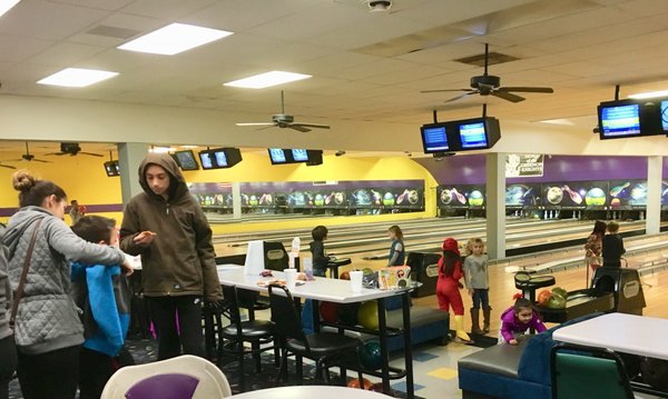 Families and children gather in a lively bowling center with lanes and overhead scoring monitors in the background.