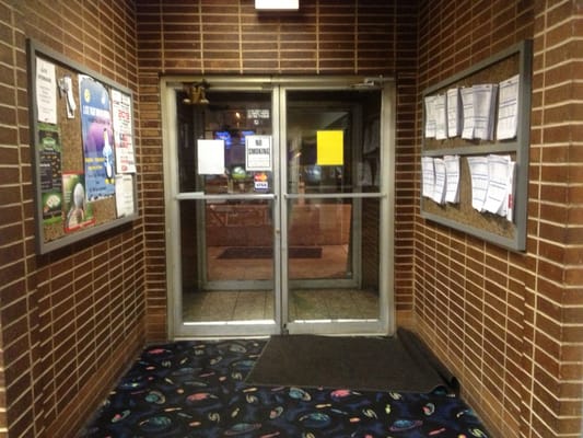 Interior entrance doors flanked by brick walls and bulletin boards covered in league standing sheets.