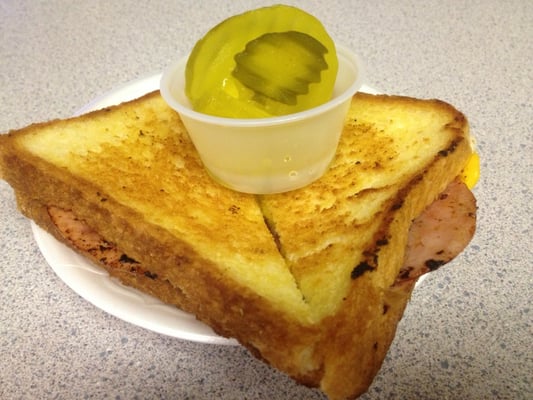 A grilled ham sandwich on thick Texas toast with a pickle slice on top, served on a white plate.