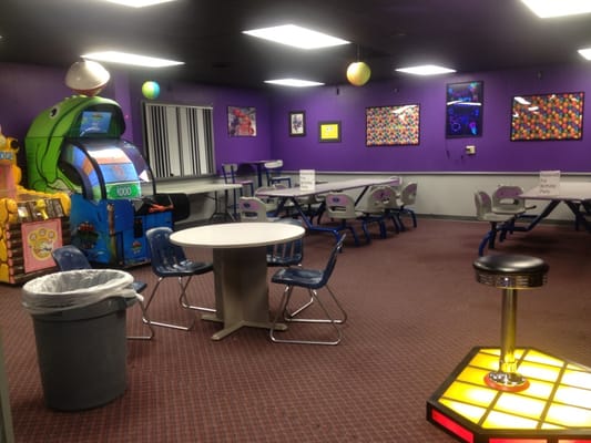 A party room with purple walls, several long tables, chairs, and arcade games in the background.