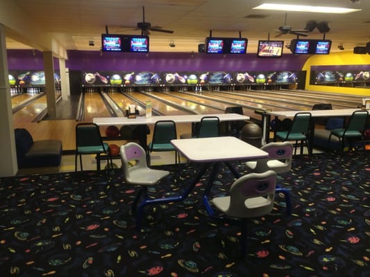 An interior view showing several bowling lanes with overhead scoring monitors and seating tables in the foreground.