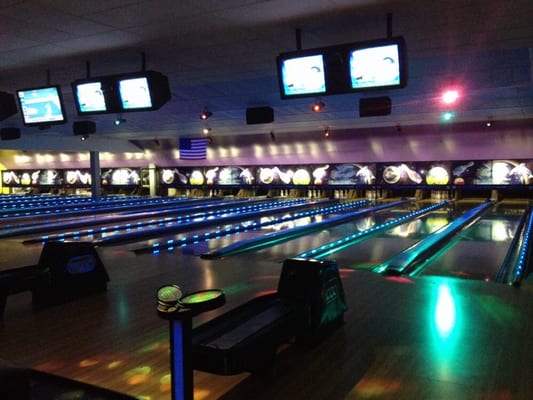 Wide view of bowling lanes under vibrant blue and green cosmic lighting with overhead scoring monitors.