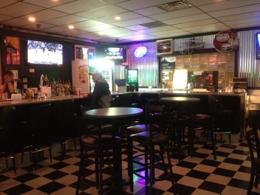 A bar and lounge area with checkered floors, high-top tables, bar stools, and televisions showing sports.
