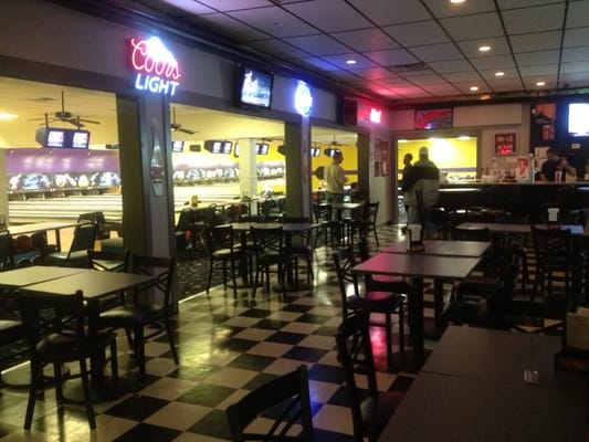 A bar and lounge area with checkered floors and tables overlooking several bowling lanes.