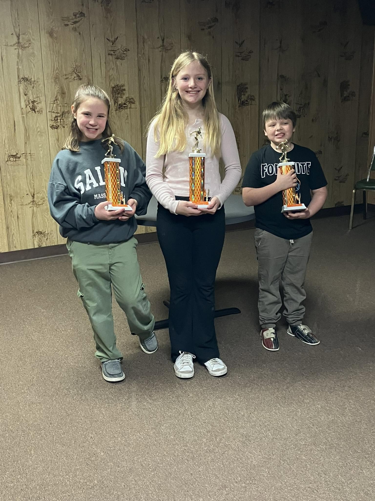 Three smiling children hold tall bowling trophies in a room with wood paneling.
