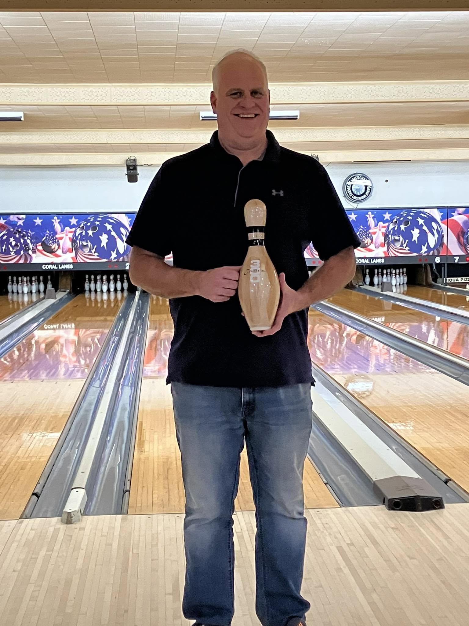 A smiling man stands on a bowling lane holding a trophy bowling pin marked with the number 300.