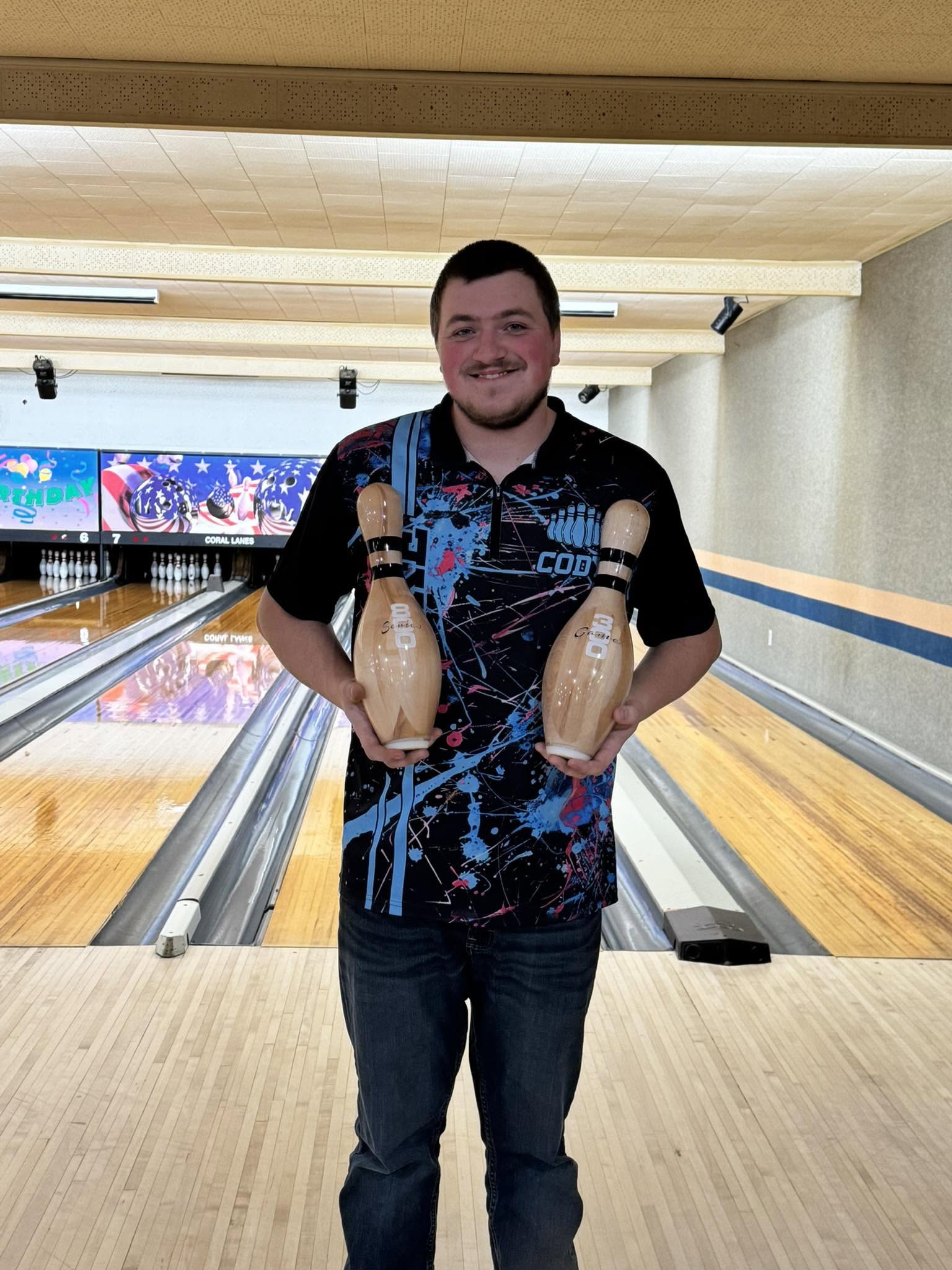 A smiling man stands on a bowling lane holding award pins for a 300 game and an 800 series.