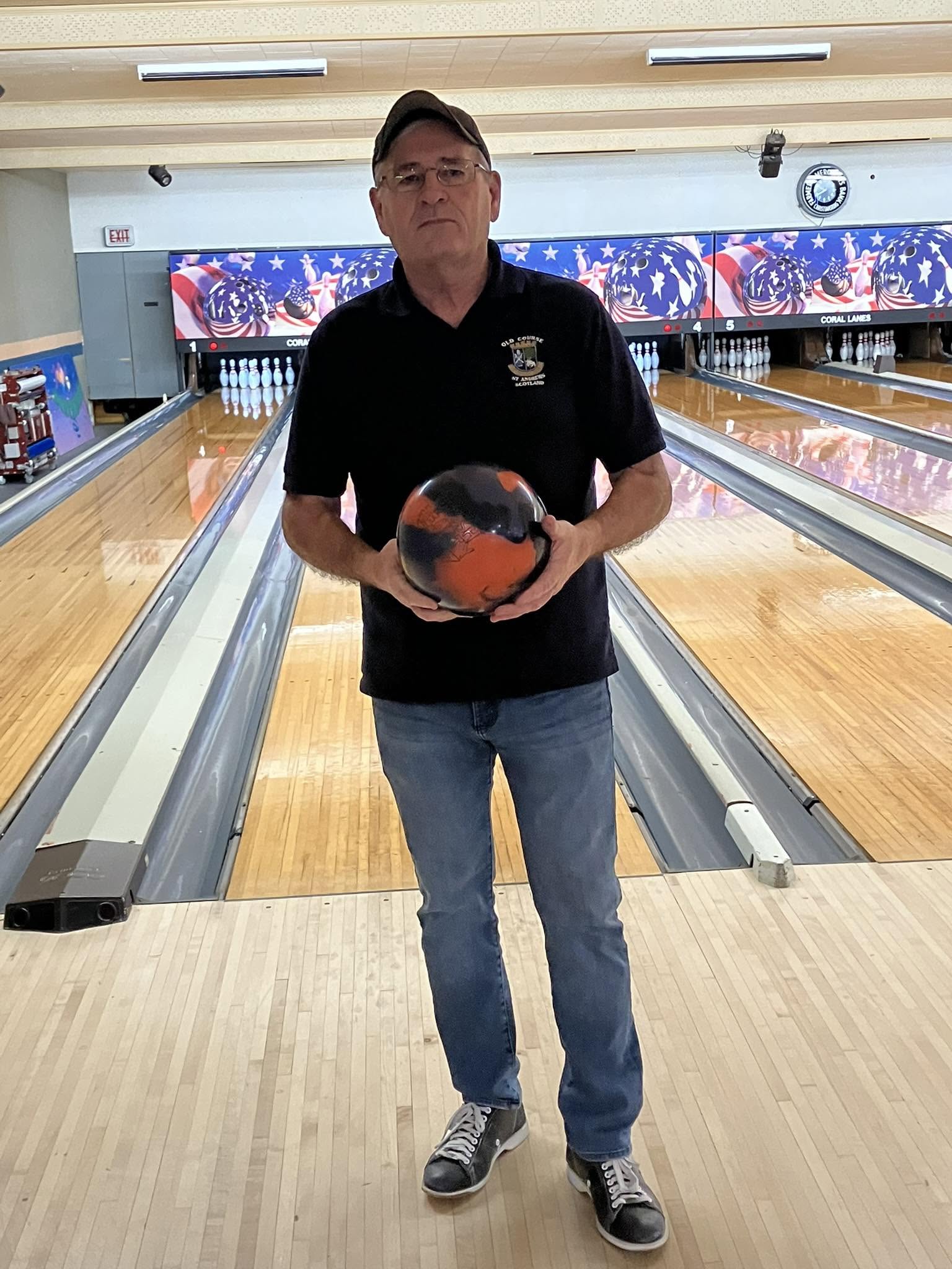 A man in a black polo and jeans stands on a bowling lane approach holding a black and orange marbled bowling ball.