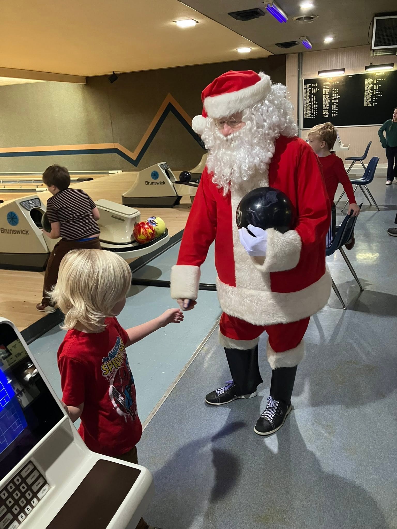 Santa Claus in a red suit and bowling shoes holds a bowling ball while talking to a young child at the lanes.