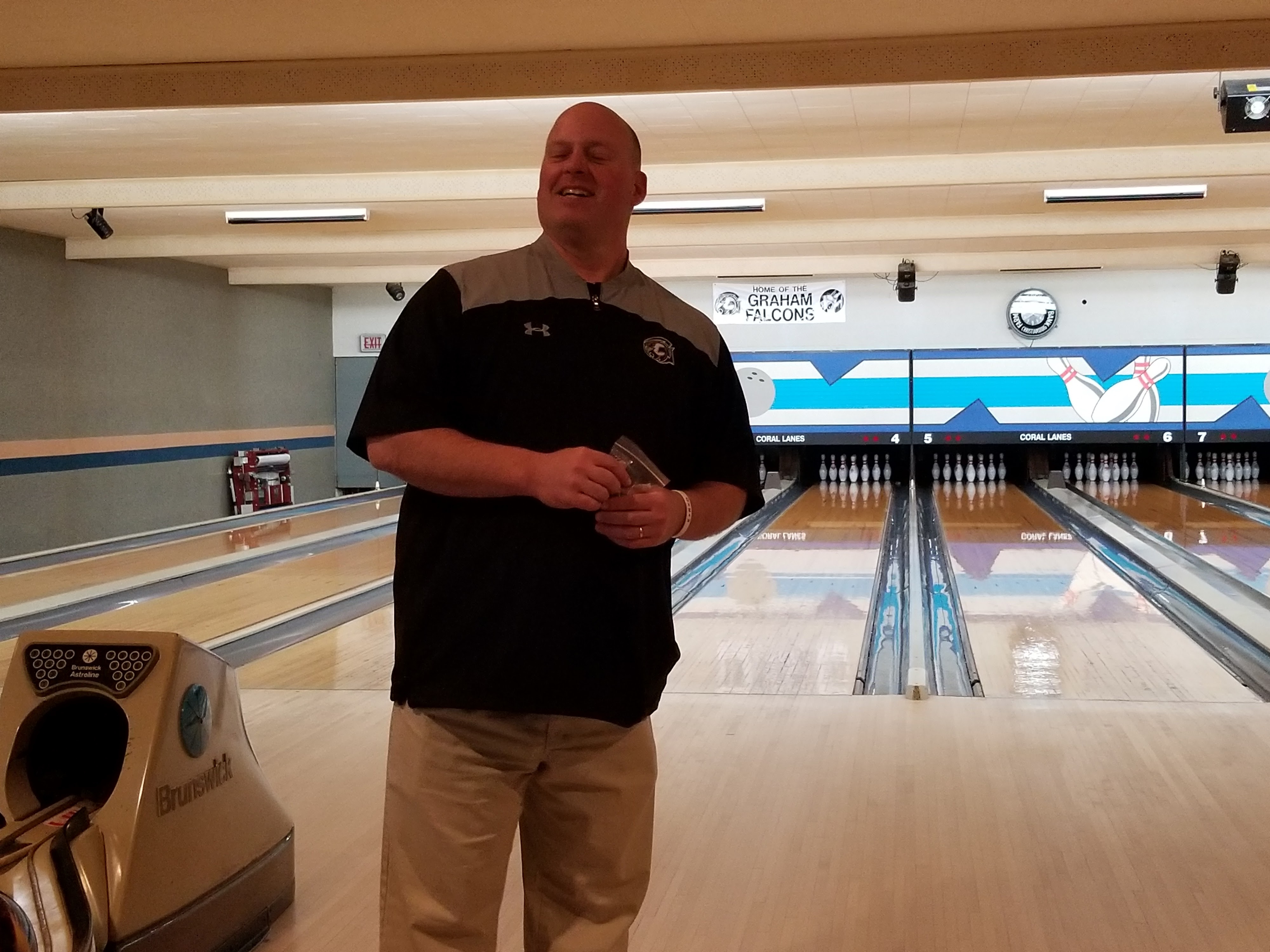 A man stands on a bowling approach in front of lanes with a Graham Falcons banner in the background.