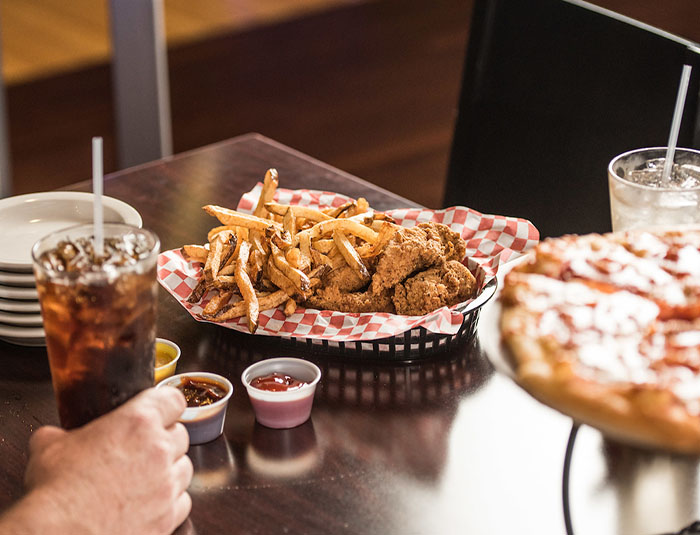 A basket of chicken tenders and fries next to a pepperoni pizza and sodas on a table.