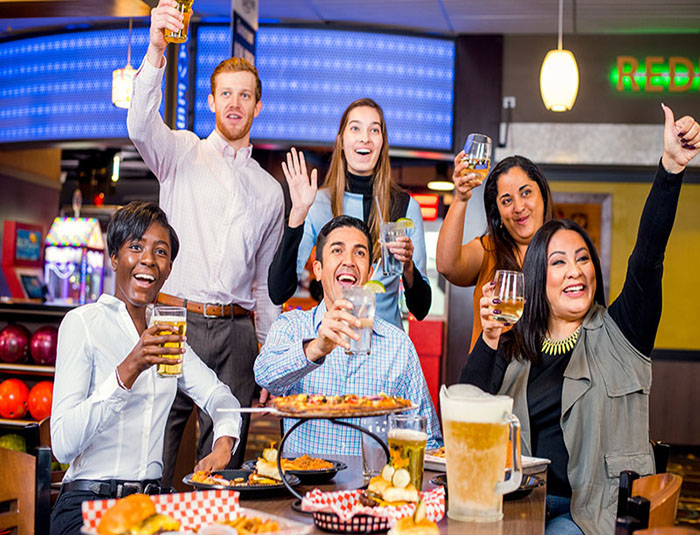 A group of friends cheering with drinks and a table full of pizza and sliders at a bowling center.