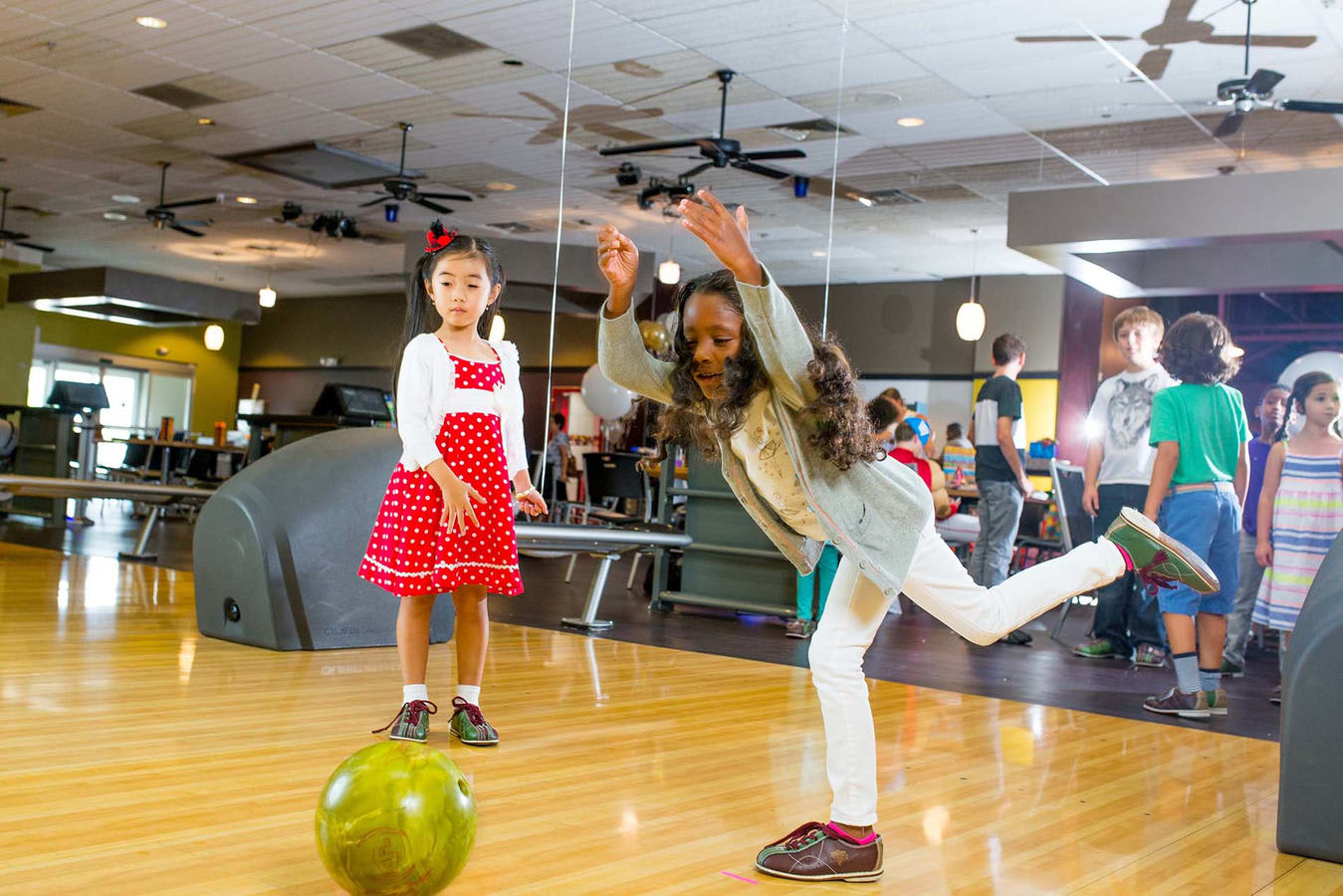 A young girl in mid-motion follows through after throwing a green bowling ball down the lane.