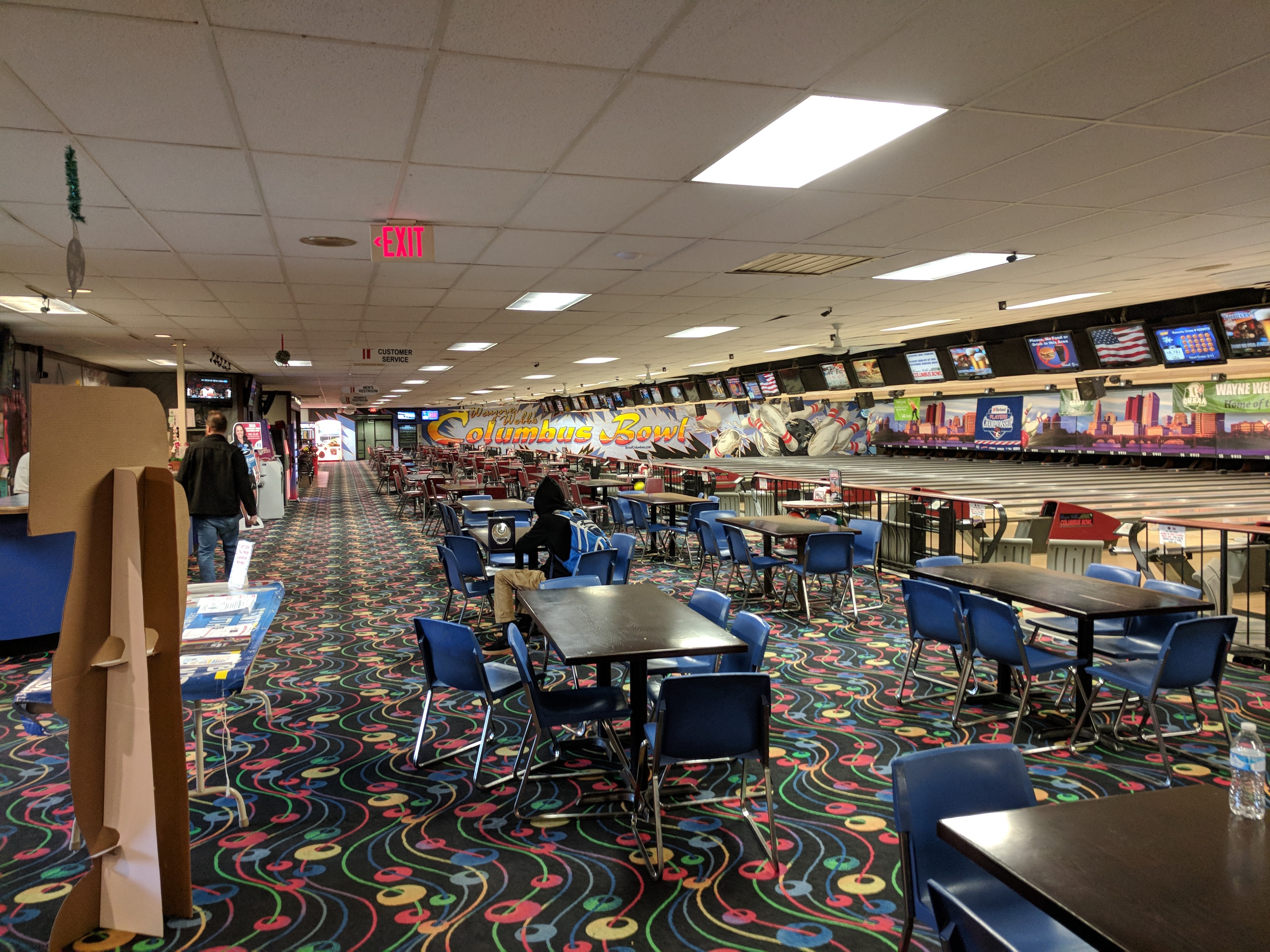 A wide interior view of a bowling center featuring many dining tables and blue chairs next to a long row of lanes.