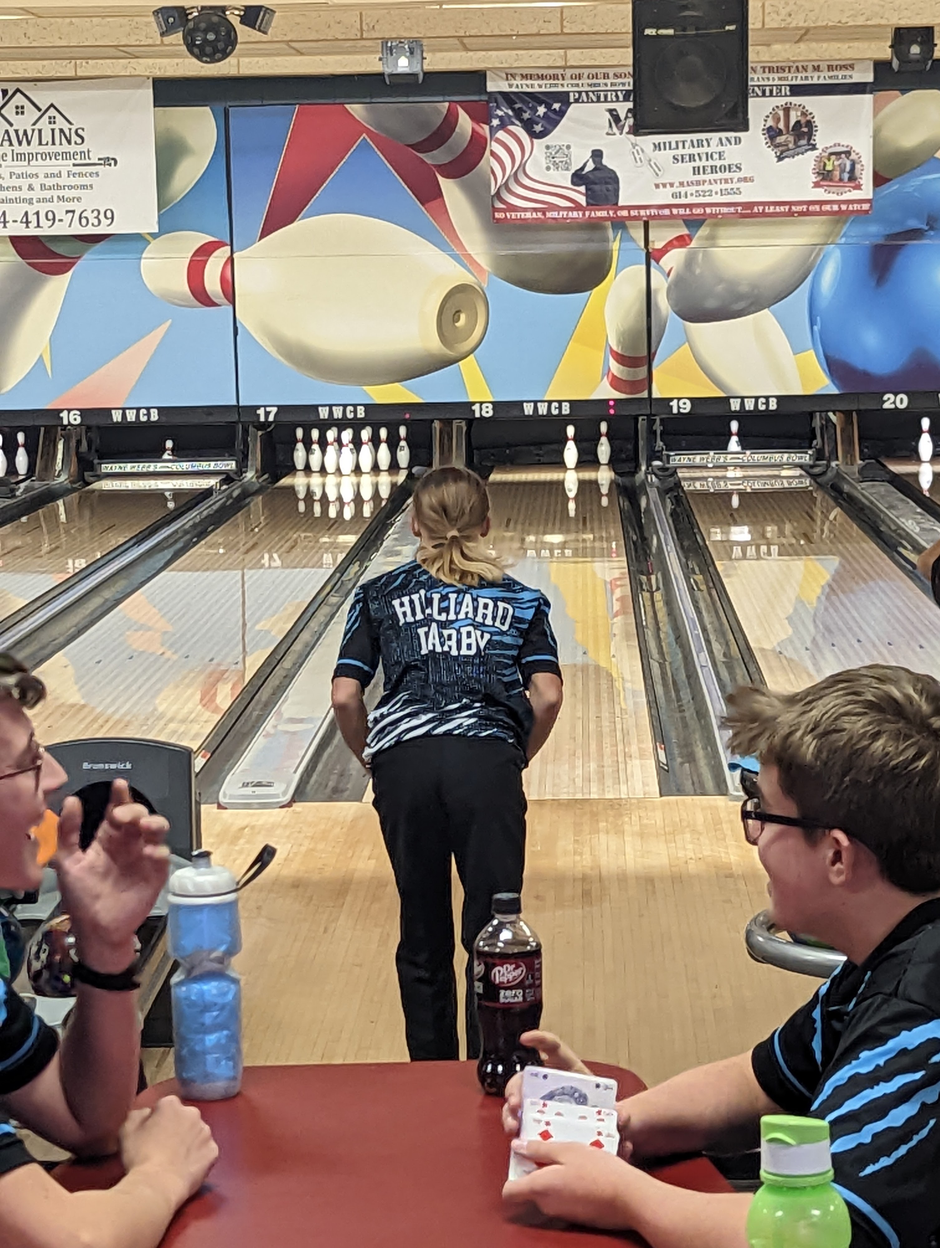 A teenager in a Hilliard Darby bowling jersey stands on the lane while friends play cards in the foreground.