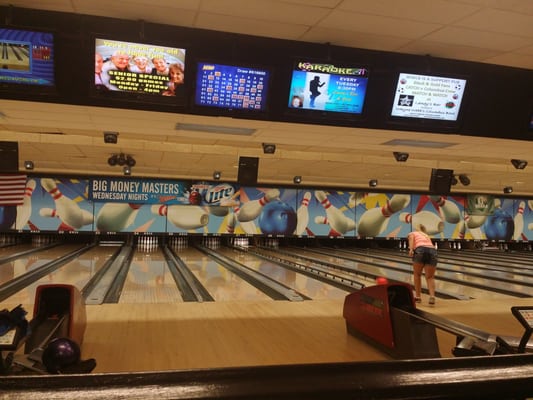 A row of bowling lanes with overhead scoring monitors and a woman preparing to bowl.