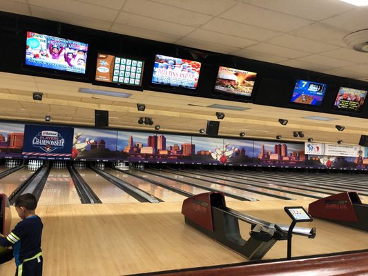 Interior view of bowling lanes with a city skyline mural and digital scoring monitors overhead.