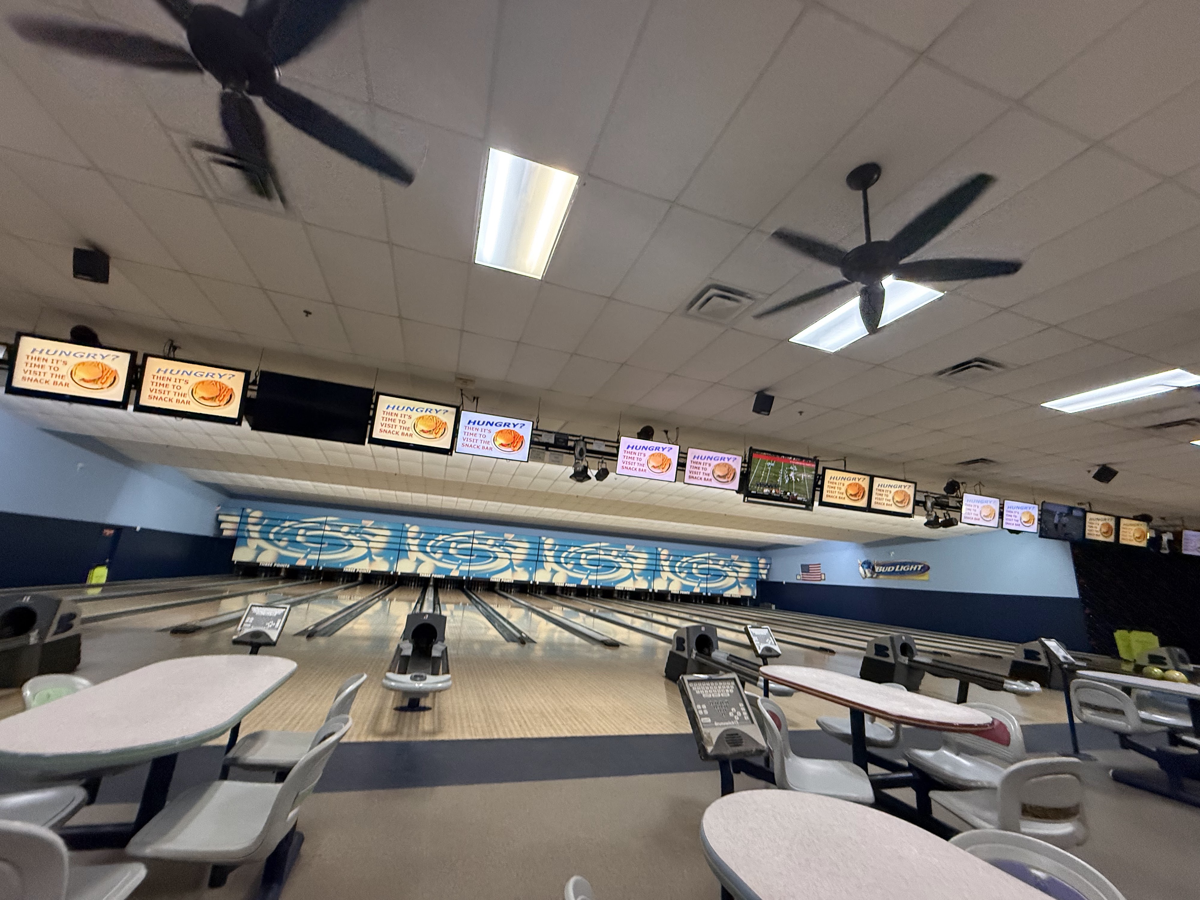 A wide interior view of bowling lanes with gray seating tables and digital scoring monitors overhead.