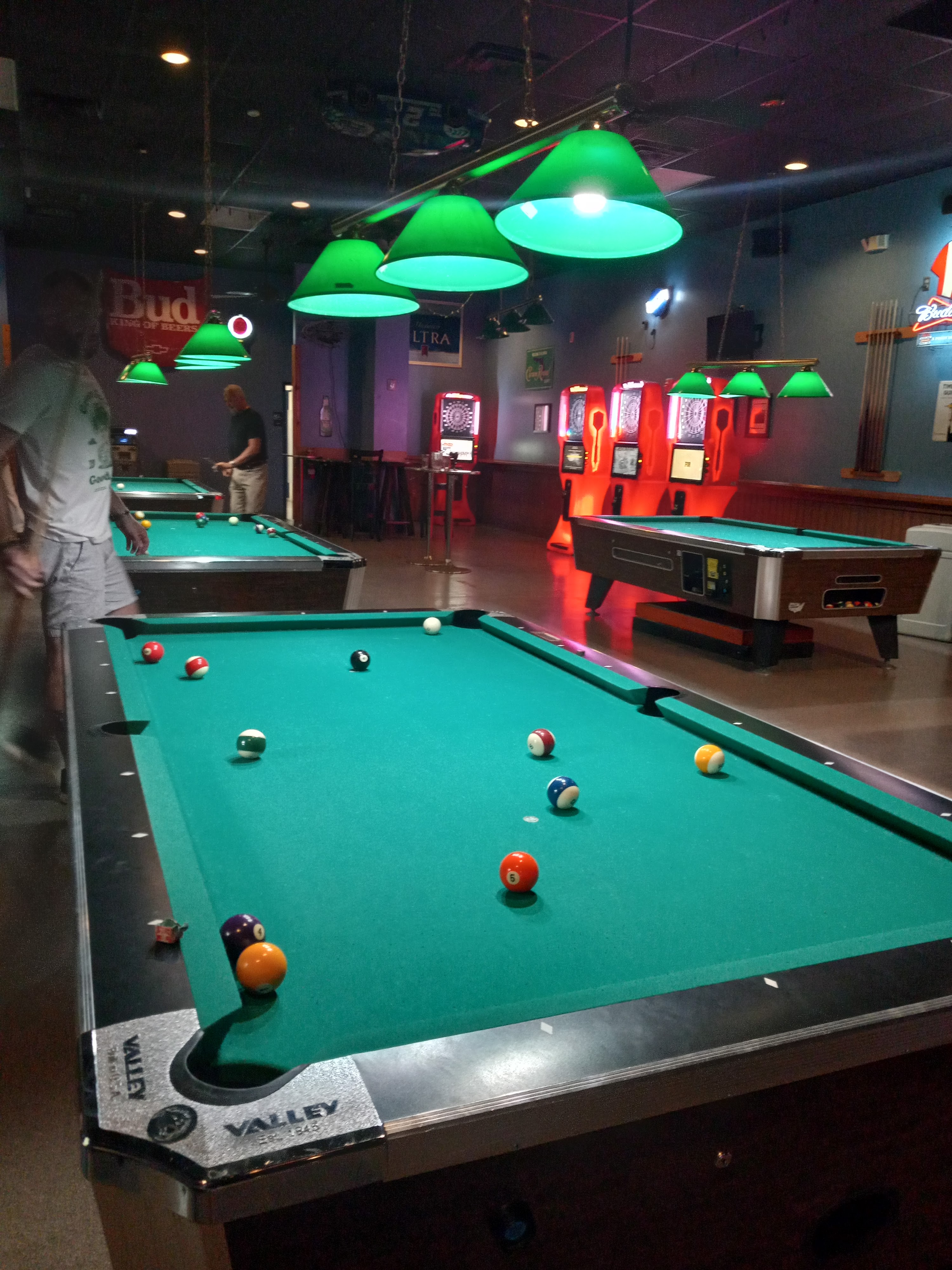Several green felt pool tables in a dimly lit billiards room with electronic dart boards in the background.
