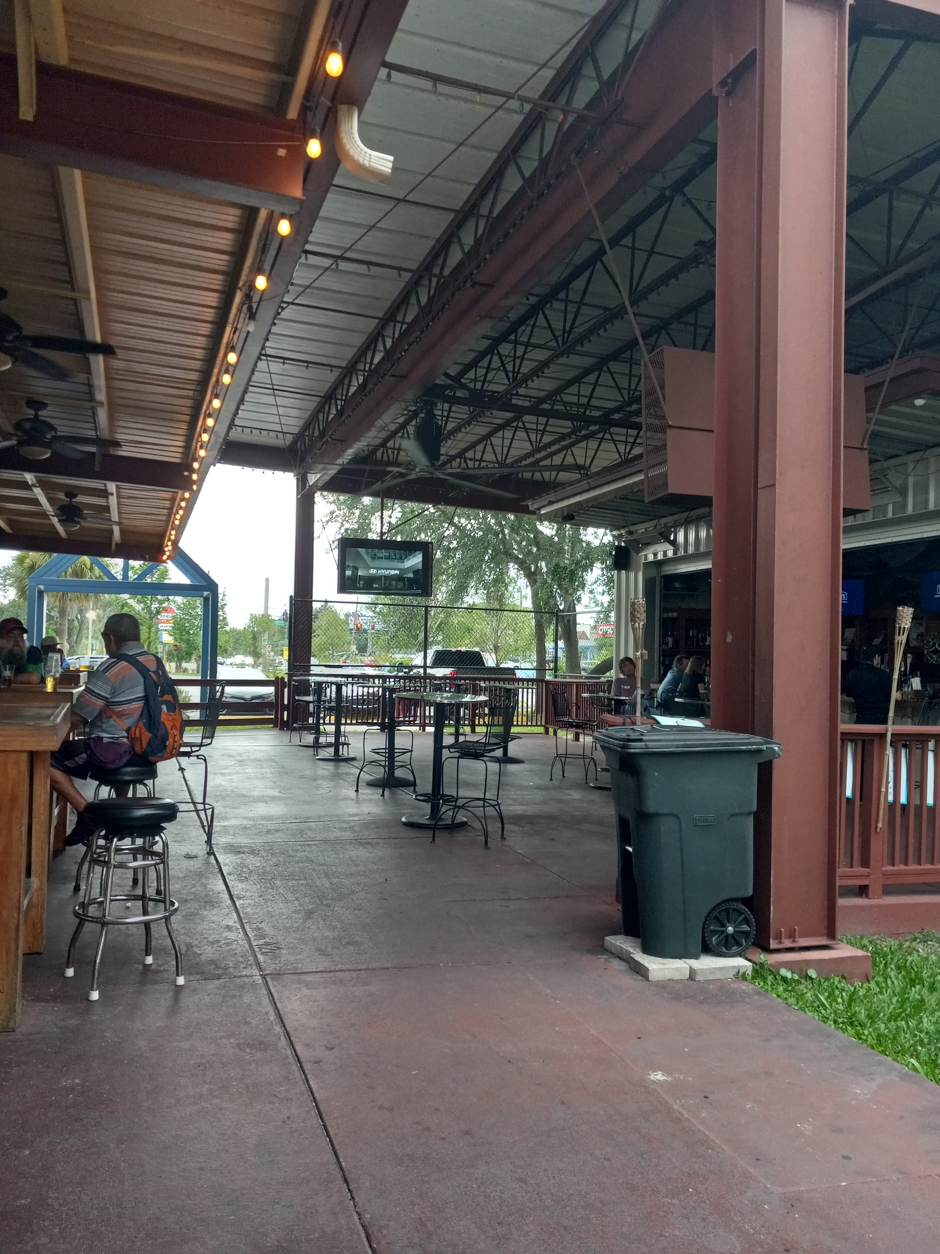 A spacious covered outdoor patio with industrial steel beams, high-top tables, bar seating, and string lights.