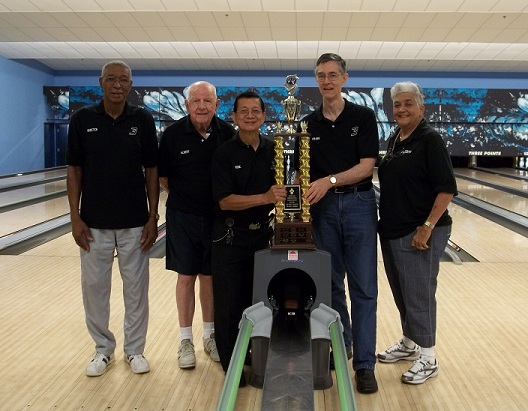 A group of five bowlers in matching black shirts posing with a large gold trophy on the lanes.