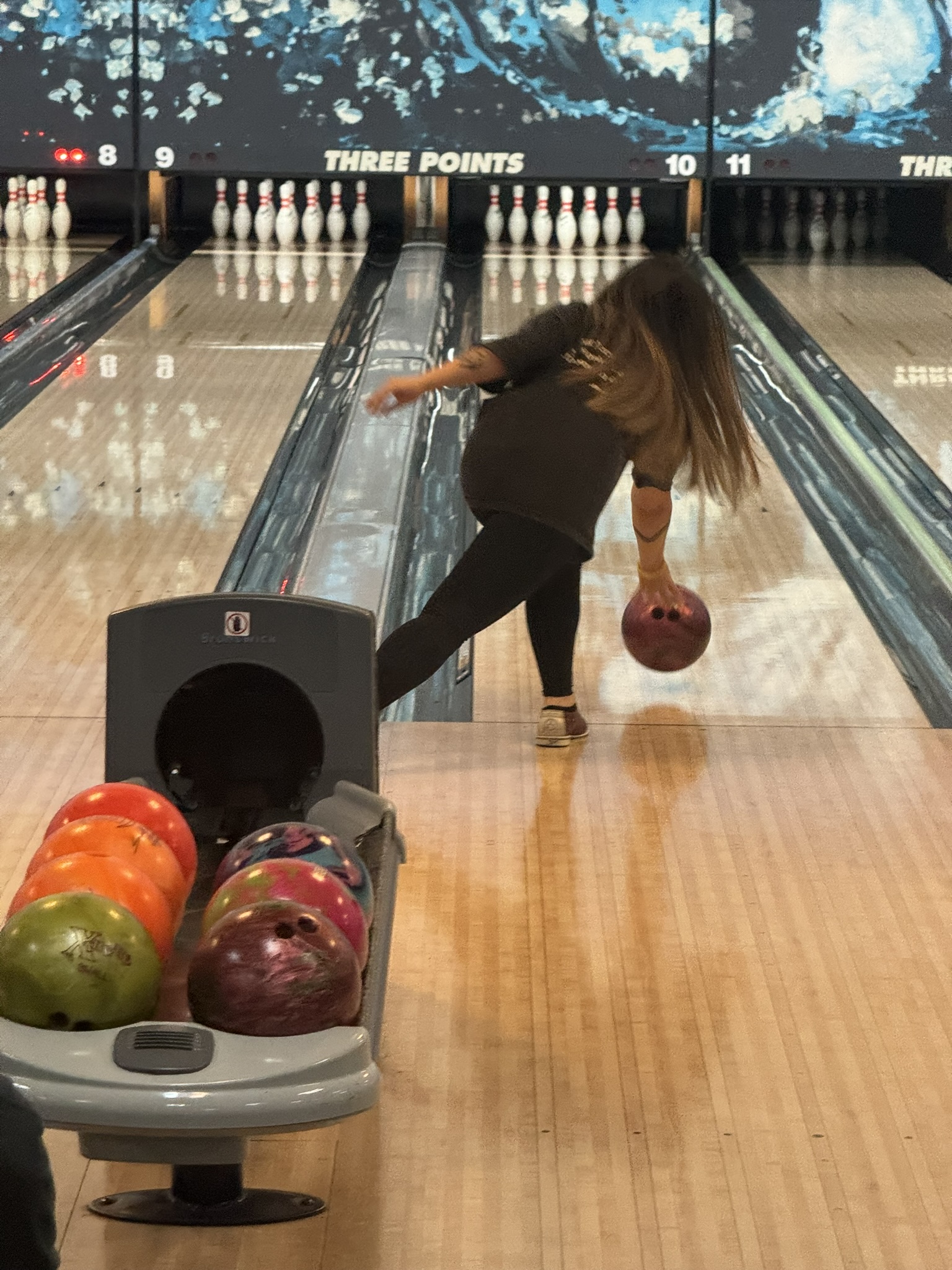A bowler in a black shirt releases a red ball down a bowling lane next to a full ball return.
