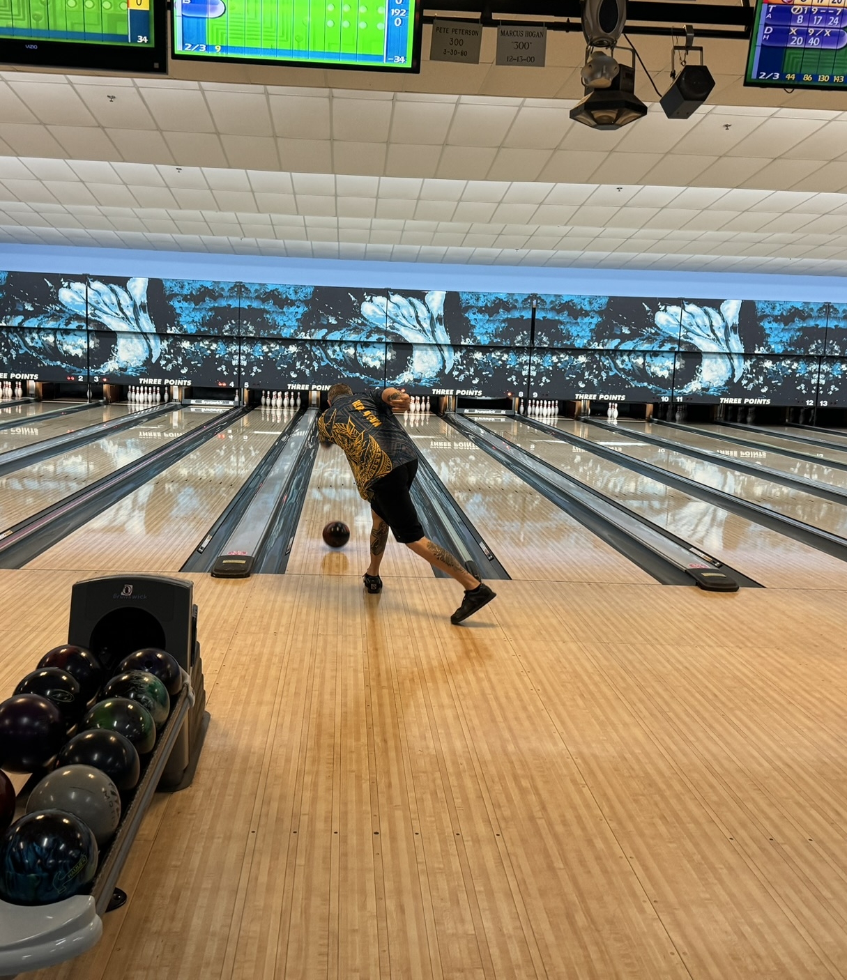 A bowler in a black and gold jersey releases a ball down a bright wooden lane under scoring monitors.