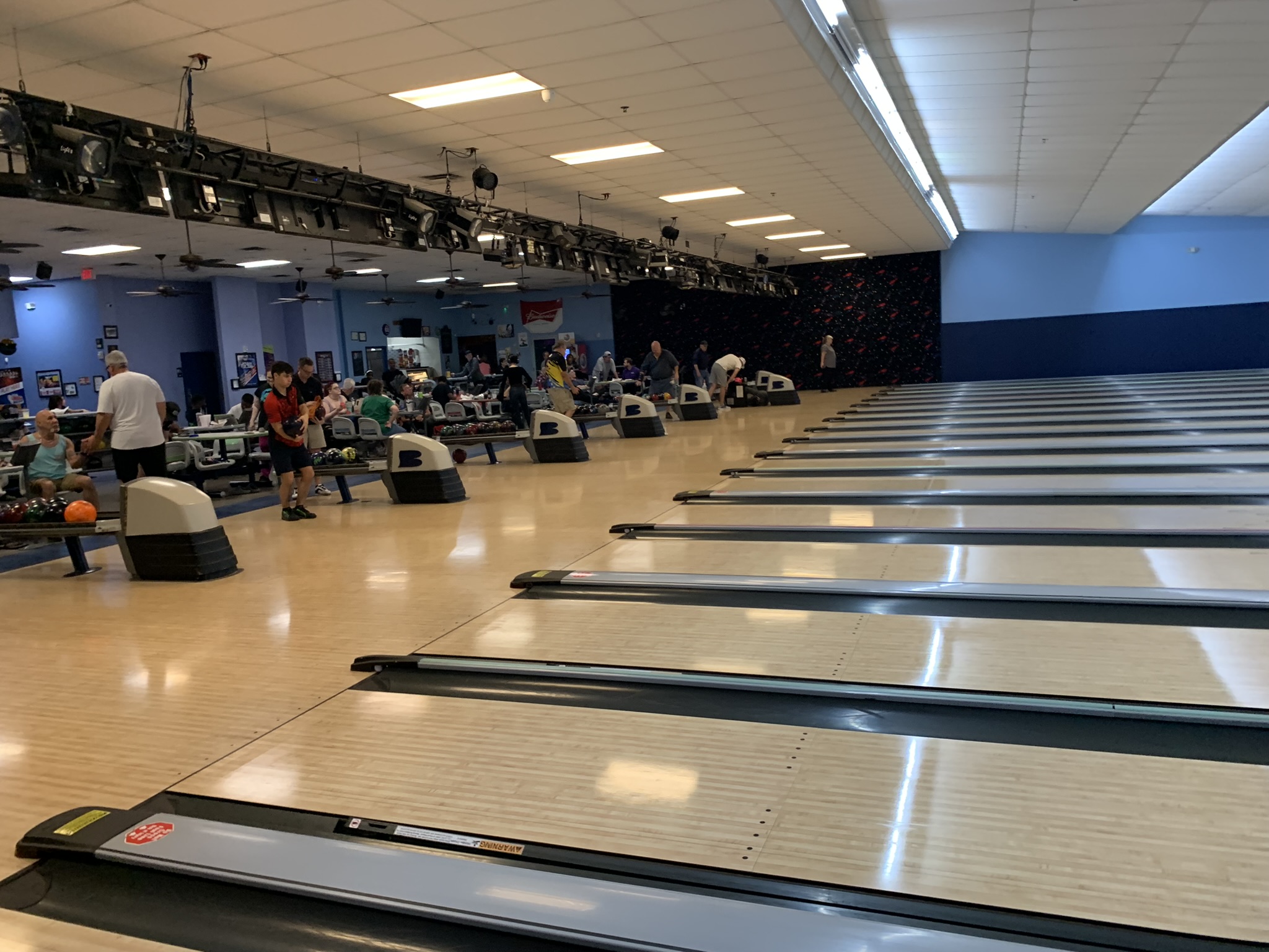 A bustling bowling alley with many people playing on multiple polished lanes under bright indoor lighting.
