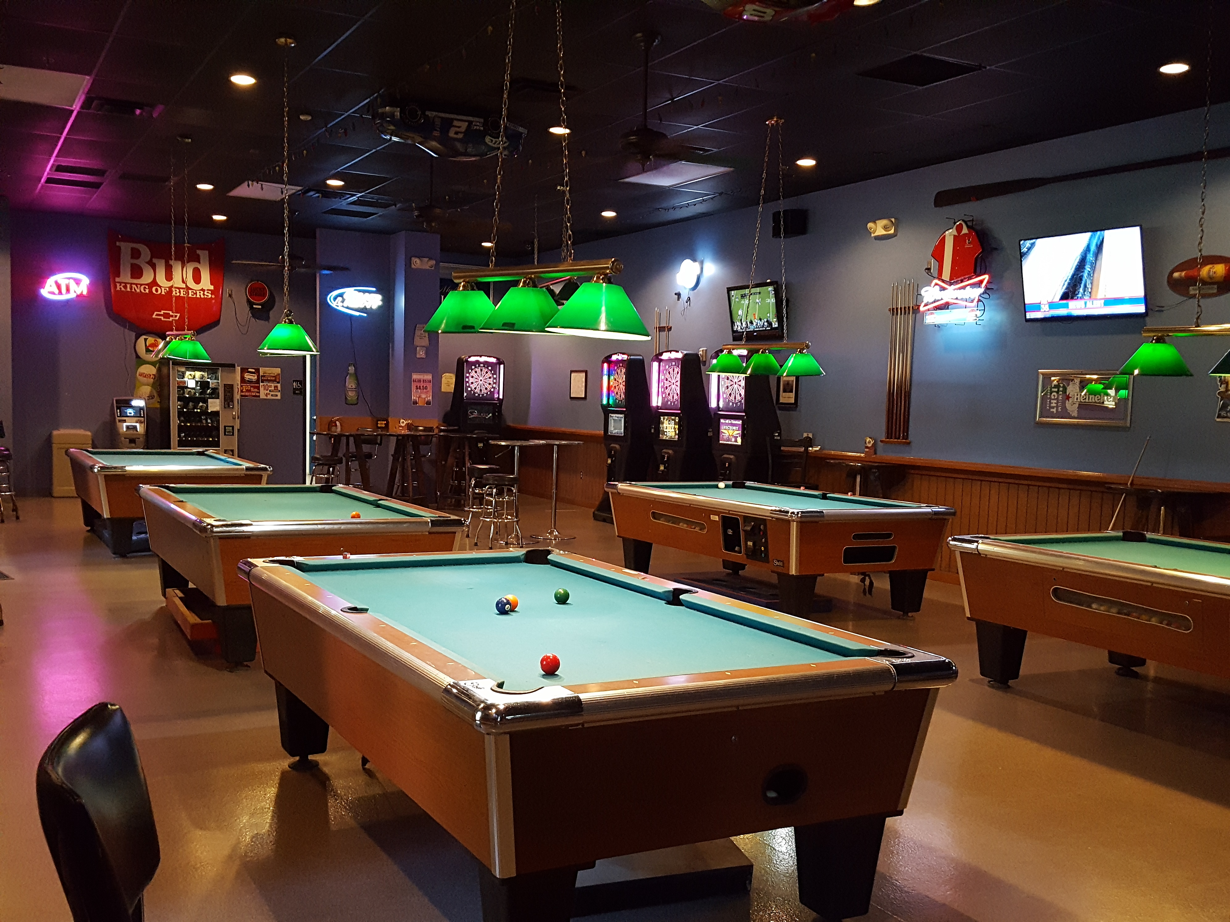 A spacious billiards room with several green-felt pool tables under hanging lamps and neon bar signs.