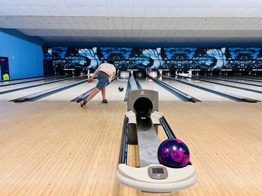 A bowler in a white shirt and shorts releases a ball down a lane in a brightly lit bowling center.