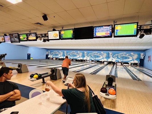 People bowling and relaxing at tables in a brightly lit bowling alley with overhead scoring monitors.